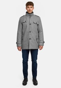 Mann steht und trägt einen grauen, geknöpften Wollmantel mit Klappentaschen, dunkle Jeans und schwarze Schuhe vor einem schlichten weißen Hintergrund.