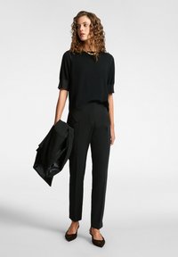 Haut en tricot noir à manches courtes, accompagné d'un pantalon noir ajusté. Le modèle tient une veste noire assortie et porte des chaussures marron texturées.
