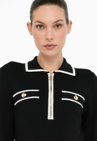 Haut en tricot noir avec bord blanc, présentant un devant zippé et deux poches décoratives avec des accents dorés. Design de col structuré.