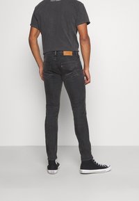 Man som bär svarta slim-fit jeans, svarta högklackade Converse-skor och en mörkgrå t-shirt med texten "NIGHT ADDICT" på ryggen.
