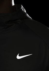 Chaqueta deportiva negra con una textura suave y mate. Logo prominente de Nike en blanco en el pecho. El diseño incluye una capucha y patrones sutiles.