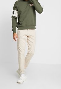 Felpa verde con maniche a contrasto bianche e toppe rettangolari; abbinata a pantaloni beige e sneakers bianchi. Materiale in cotone misto resistente.