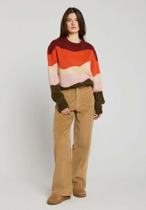 Jeune femme aux cheveux bruns et lisses porte un pull en maille multicolore, un pantalon large en velours beige et des chaussures en daim beige à enfiler, debout contre un mur uni.