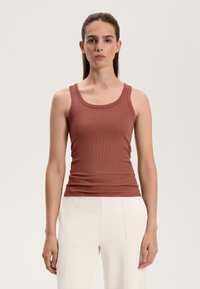 Fitted ribbed tank top in rust color with wide shoulder straps and a round neckline, paired with cream-colored trousers.