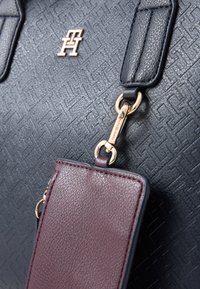 Fekete, textúrált bőrtáska aranyozott hardverrel, domináló logóval, valamint egy levehető, sima felületű bordó pénztárral rendelkezik.