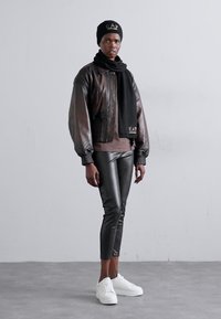 Giacca bomber in pelle marrone, leggings neri aderenti, sneaker bianche, berretto nero e sciarpa con logo. Texture morbide e vestibilità su misura.