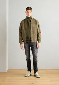 Maslinazeleni zamak bomber, rebrasti turtleneck, tamni traperice i tenisice sa zelenim prugama; model stoji naslonjen na svijetlosivu zid.
