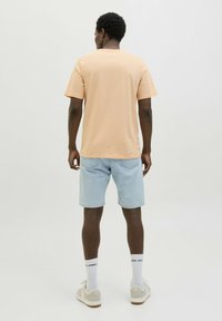 Camiseta de algodón color durazno claro con mangas cortas, combinada con pantalones cortos de mezclilla azul claro. Lleva calcetines blancos y zapatillas beige.