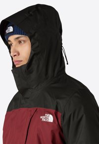 Giacca nera e bordeaux con cappuccio, dotata di chiusura con zip e logo sul petto, realizzata in un materiale liscio e resistente alle intemperie.
