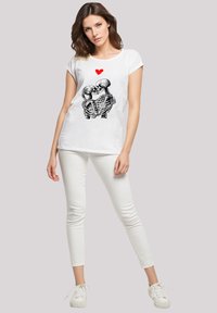 Weißes Baumwoll-T-Shirt mit einem Grafikmotiv von zwei Skeletten und einem roten Herzen darüber, kombiniert mit weißen Skinny-Jeans und weißen Sneakers.