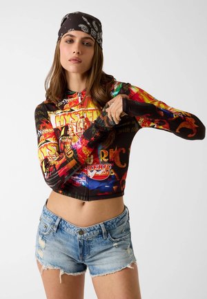 Giovane donna con un top corto a maniche lunghe colorato e grafico, shorts di denim strappati e un bandana nero, che si sistema la manica.