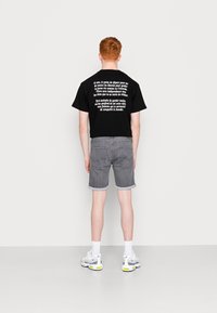 Camiseta negra con texto blanco en la parte posterior, combinada con pantalones cortos de mezclilla grises. El modelo lleva zapatillas grises y blancas, de pie contra un fondo claro.