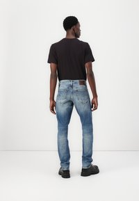 Egy férfi fekete pólóban, fakó kék farmernadrágban és fekete cipőben áll, háttal a kamerának, egy fehér háttér előtt.