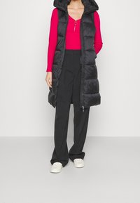 Czarna puchowa kamizelka z kapturem, zapinana na zamek, z bocznymi kieszeniami, na jasnoróżowej bluzce z długim rękawem i czarnych szerokich spodniach. Białe trampki.