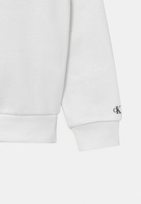 Biała bluza z miękkiej tkaniny, z prążkowanymi mankietami i małym czarnym logo na rękawie. Prosty, klasyczny design bez wzorów.