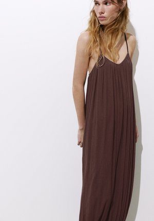 Femme aux longs cheveux blonds portant une robe longue plissée marron sans manches avec des fines bretelles, debout devant un fond blanc uni.