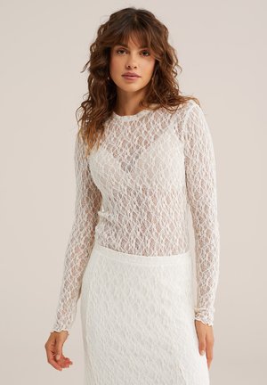 Žena s vlnitými hnedými vlasmi, oblečená v bielom čipkovanom topaní s dlhým rukávom a zodpovedajúcej bielej čipkovej sukni na neutrálnej pozadí.