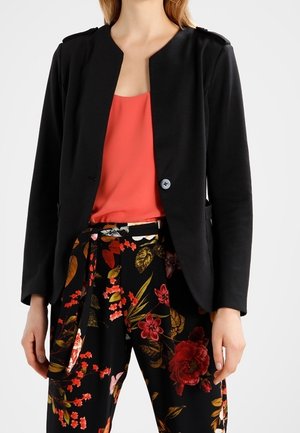Woman wearing a black single-button blazer over a coral blouse and black pants with red, orange, and yellow floral patterns.