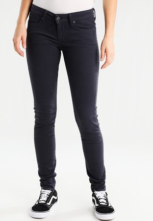 Persona vistiendo jeans azul marino oscuros de corte ajustado y zapatillas blancas y negras, de pie frente a un fondo blanco liso.