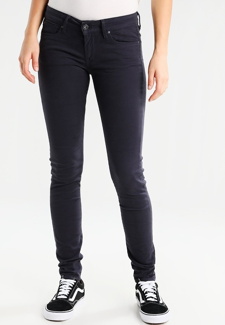 Personne portant un jean slim bleu marine foncé et des baskets noires et blanches, debout devant un fond blanc uni.