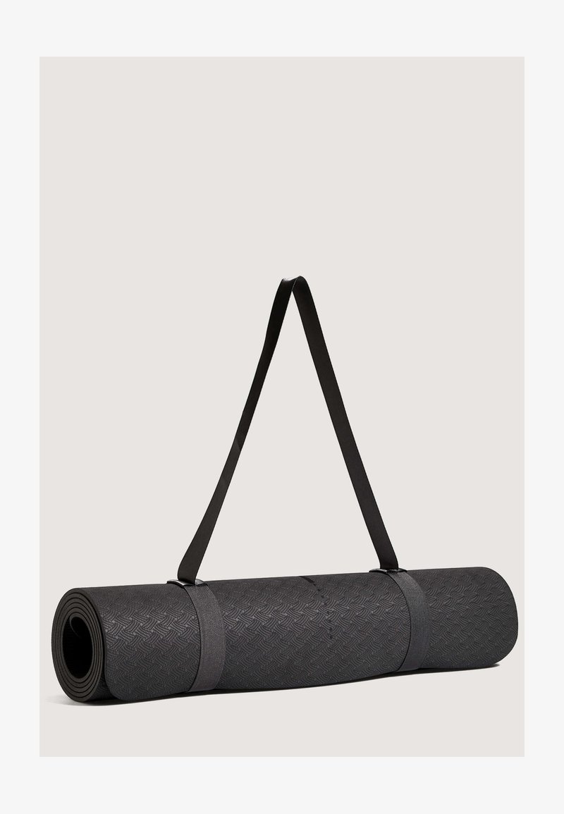 Musta joogamatto rullattuna, valmistettu teksturoidusta materiaalista, kiinnitetty harmailla kantohihnoilla. Pinnassa on geometrinen kuvio.