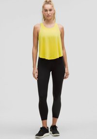 Geel mouwloos topje gecombineerd met zwarte strakke leggings en zwarte sneakers. Het topje heeft een losse, vloeiende snit.