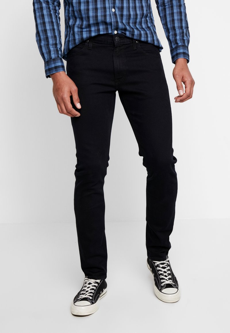 Jean skinny noir à la texture lisse, associé à une chemise à carreaux bleue et noire et des baskets noires. Accent sur l'ajustement des jambes et le tissu.