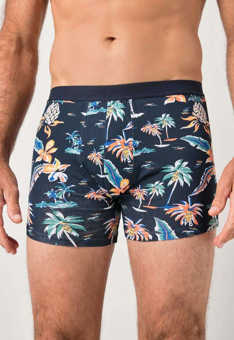 Uomo che indossa boxer blu navy con stampa tropicale di fiori e palme, in piedi contro uno sfondo semplice.