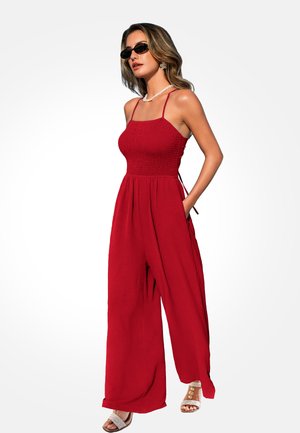 Donna che indossa una tuta rossa senza maniche con gambe larghe, occhiali da sole, sandali bianchi, collana di perle e orecchini a forma di fiore.