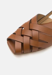 Sandal din piele maro cu vârf deschis, având un design cu curea împletită, textură netedă și o talpă plată. Accentuată cu margini întunecate și cusături minime.