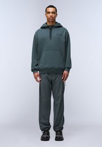 Felpa con cappuccio verde scuro, dotata di tasca frontale, abbinata a pantaloni larghi dello stesso colore. Scarpe nere con lacci completano l'outfit.
