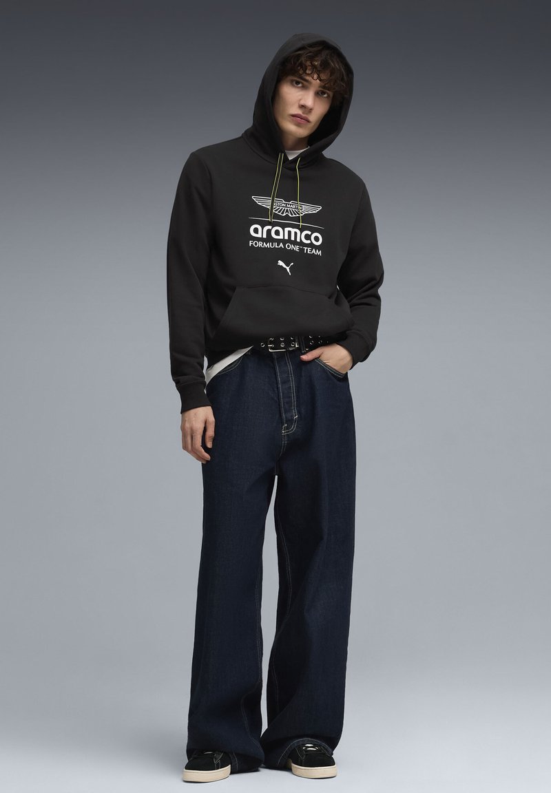 Sweat à capuche noir avec logo et cordon de serrage, associé à un jean en denim foncé surdimensionné. Le modèle porte des chaussures noires avec une semelle crème.