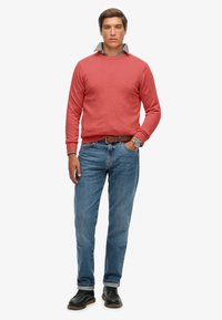 Rode katoenen trui over een geruit shirt, gecombineerd met blauwe denim jeans en zwarte schoenen. De outfit heeft een casual pasvorm en een gestructureerde stof.