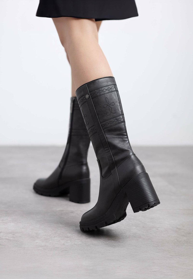 Botas de cuero negras hasta la rodilla con tacón grueso, patrones de costura intrincados y cremalleras laterales, con una suela texturizada para mayor agarre.