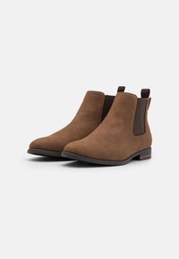 Bottines en daim marron avec panneaux latéraux élastiques, bout rond, surface texturée et petit talon avec un accent rouge contrastant.