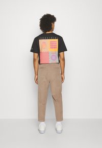 Uomo con capelli ricci che indossa una t-shirt nera con grafica, pantaloni beige, calze bianche e sneaker bianche, in piedi di spalle contro un muro semplice.