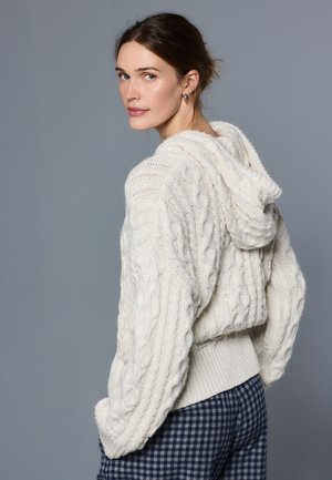 Kobieta o brązowych włosach, ubrana w biały sweter z kapturem w splocie warkoczowym oraz niebieskie spodnie w kratę, spogląda przez ramię na tle szarego tła.