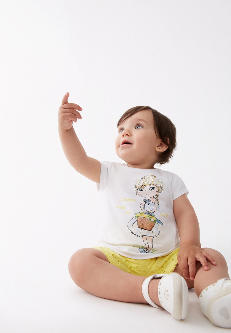Camisa blanca de manga corta con un gráfico de una chica de dibujos animados y el texto "Los limones me hacen sonreír". Combinada con pantalones cortos amarillos, sentada sobre un fondo claro.