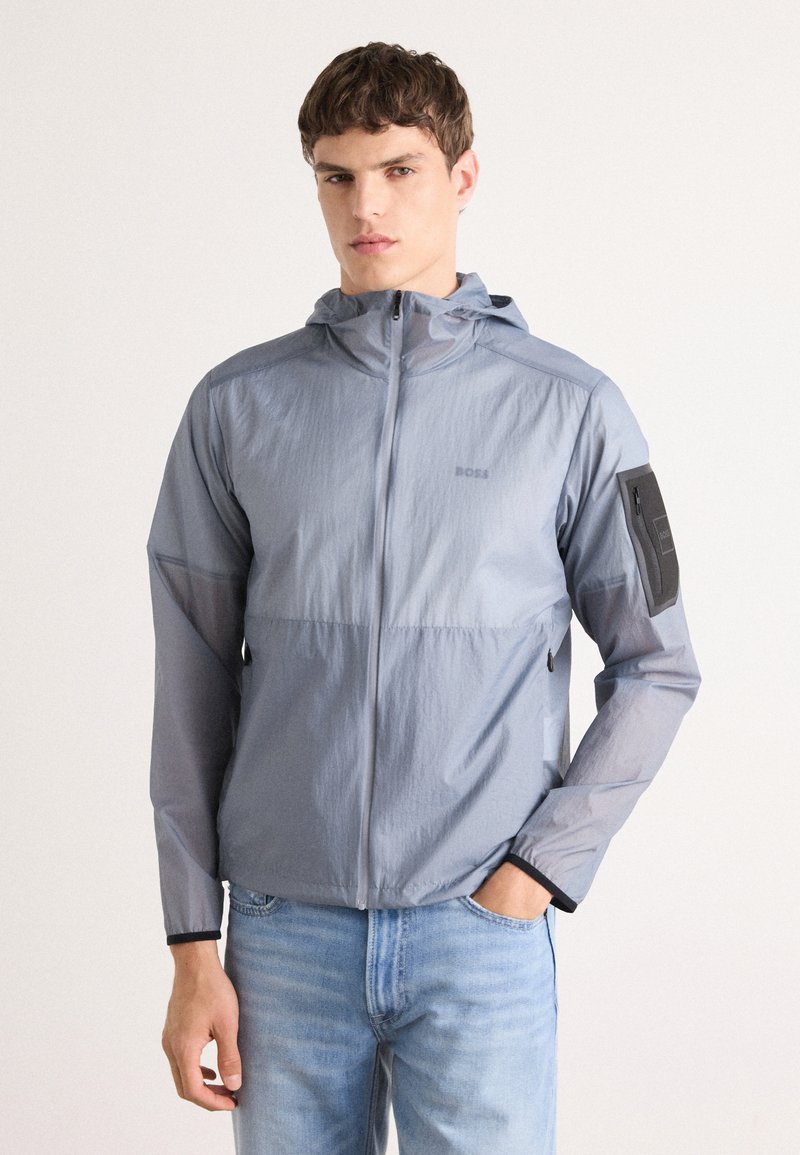 Lightweight, hooded jacket in light gray fabric. Features zip closure, elastic cuffs, and a black logo patch on the upper arm. Casual design.