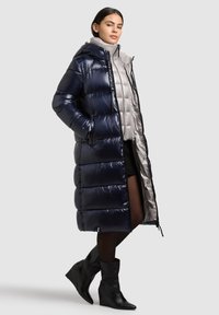 Hosszú tengerészkék pufferkabát fényes felülettel, kapucnival és steppelt mintával, világosszürke mellény fölött rétegezve. A fekete ék sarkú bokacsizma teszi teljessé a megjelenést.