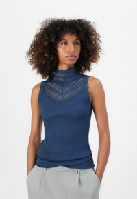 Mulher com cabelo encaracolado, vestindo uma blusa de renda azul-marinho sem mangas e calças cinza-claro com riscas finas, olhando ligeiramente para o lado com as mãos nos bolsos.