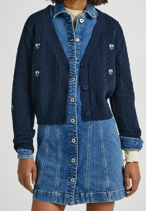 Persoon draagt een marineblauwe gebreide cardigan met kleine bloemmotieven over een denimjurk met lange mouwen en knopen.