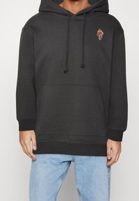 Felpa nera con cappuccio, dotata di tasca frontale, coulisse regolabile e un dettaglio di rosa ricamata colorata sul lato sinistro del petto.