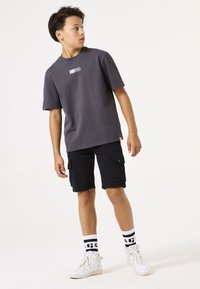 Graues T-Shirt mit kurzen Ärmeln, das ein kleines weißes Logo trägt; kombiniert mit schwarzen Cargo-Shorts und weißen High-Top-Sneakern mit schwarz gestreiften Socken.