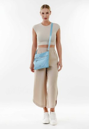 Sac bandoulière bleu clair en matériau doux, avec une poche zippée à l'avant et une longue sangle ajustable. Associé à un pantalon large beige.