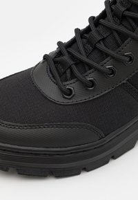 Zapatilla deportiva negra con un upper de tela texturada, acentos de goma y una suela de tracción gruesa. Los cordones y los ojales también son negros.