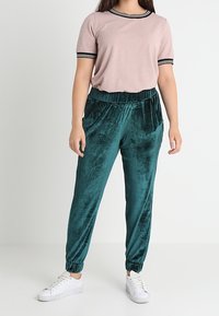 Haut rose à manches courtes avec des accents noirs, associé à un jogging en velours teal. Des baskets blanches complètent la tenue. Texture douce et coupe décontractée.