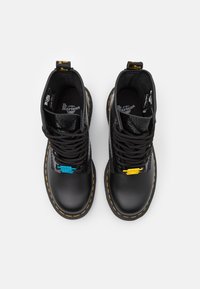 Botas de tobillo de cuero negro con costuras amarillas en contraste, paneles texturizados y cordones coloridos en azul y amarillo. Marca Dr. Martens visible.