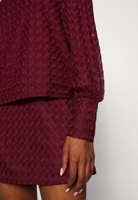 Abito bordeaux in tessuto a trama testurizzata con motivo a diamante, dotato di maniche lunghe e design a strati. Primo piano di una mano contro il tessuto.