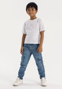 Camiseta blanca de algodón con texto bordado en verde, combinada con pantalones cargo de mezclilla azul con bolsillos laterales, complementada con zapatillas blancas.